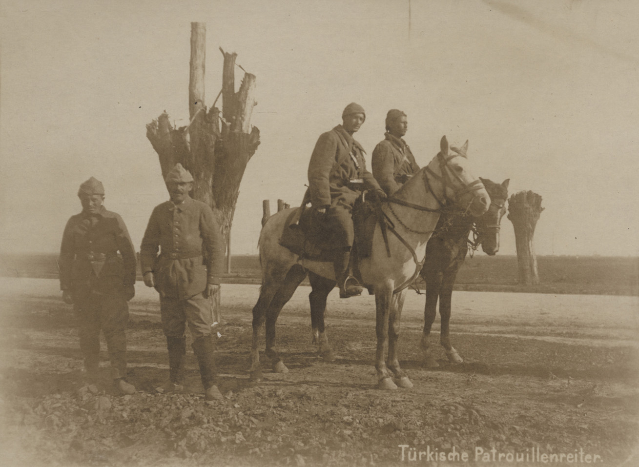 Turkish soldiers posing for a photograph in an outdoor field or plain. Two are on horses and two are standing on the ground.