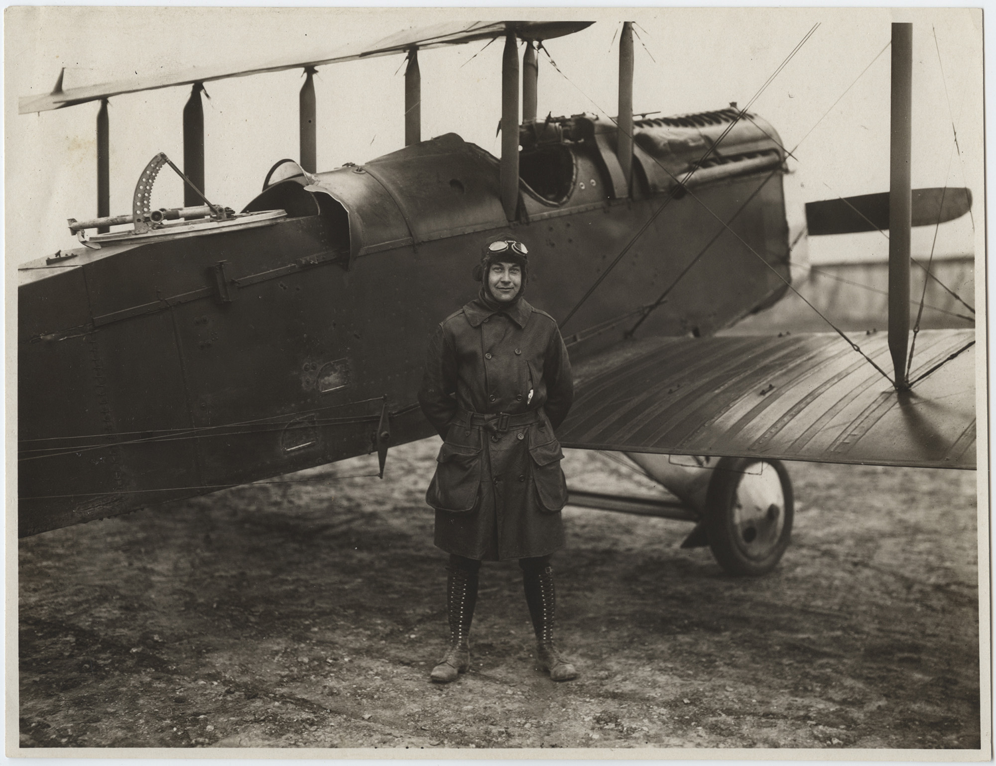 Photographie en noir et blanc d'un homme portant un trench-coat, de hautes bottes à lacets, une casquette en cuir et des lunettes de protection, debout devant un biplan de la Première Guerre mondiale
