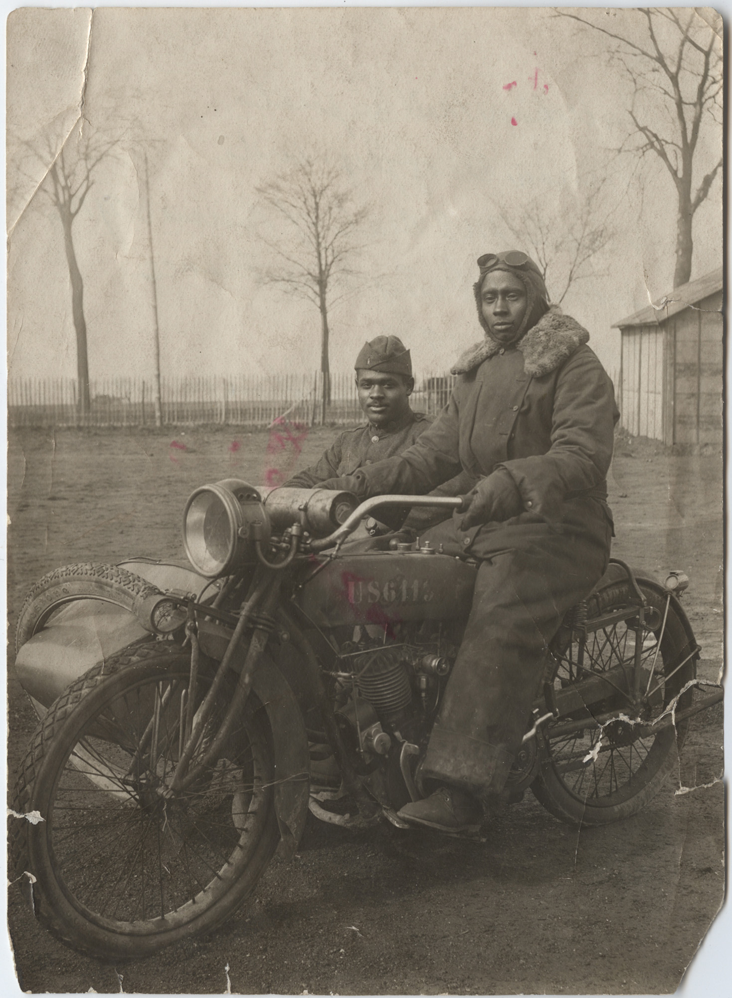 Photographie en noir et blanc d'un homme noir vêtu d'une combinaison épaisse à col en fourrure, d'une casquette en cuir et de lunettes de protection, assis sur une moto. Un autre homme noir en uniforme ordinaire est assis dans le side-car.