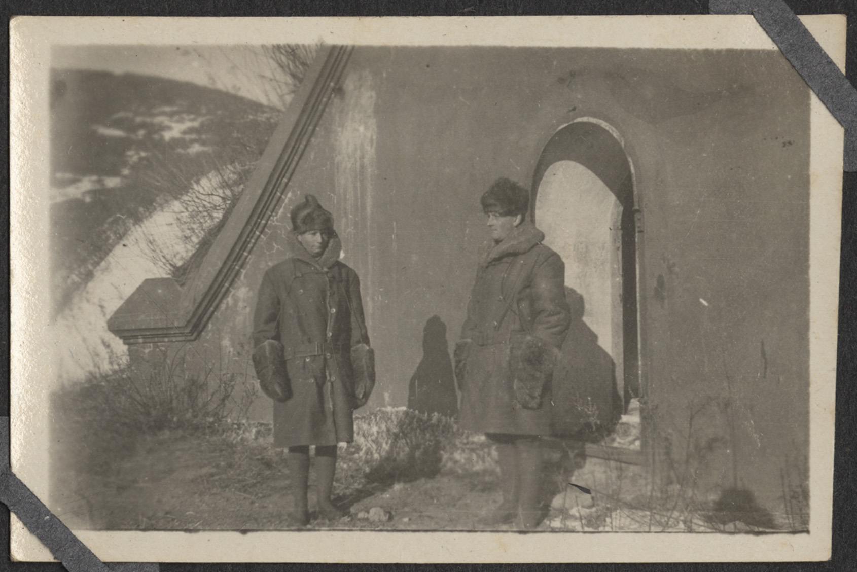 Photographie en noir et blanc de deux hommes portant des manteaux épais, des chapeaux, des bottes et des gants, debout dehors sous le soleil d'hiver