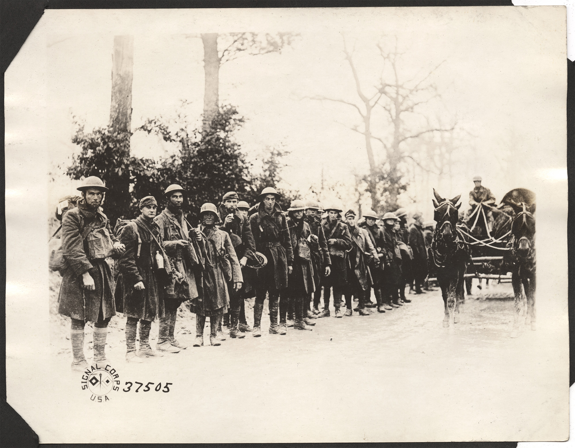 Photographie en noir et blanc d'un grand groupe de soldats alignés près d'une route et d'une charrette tirée par des chevaux. Ils portent des imperméables et des chapeaux.