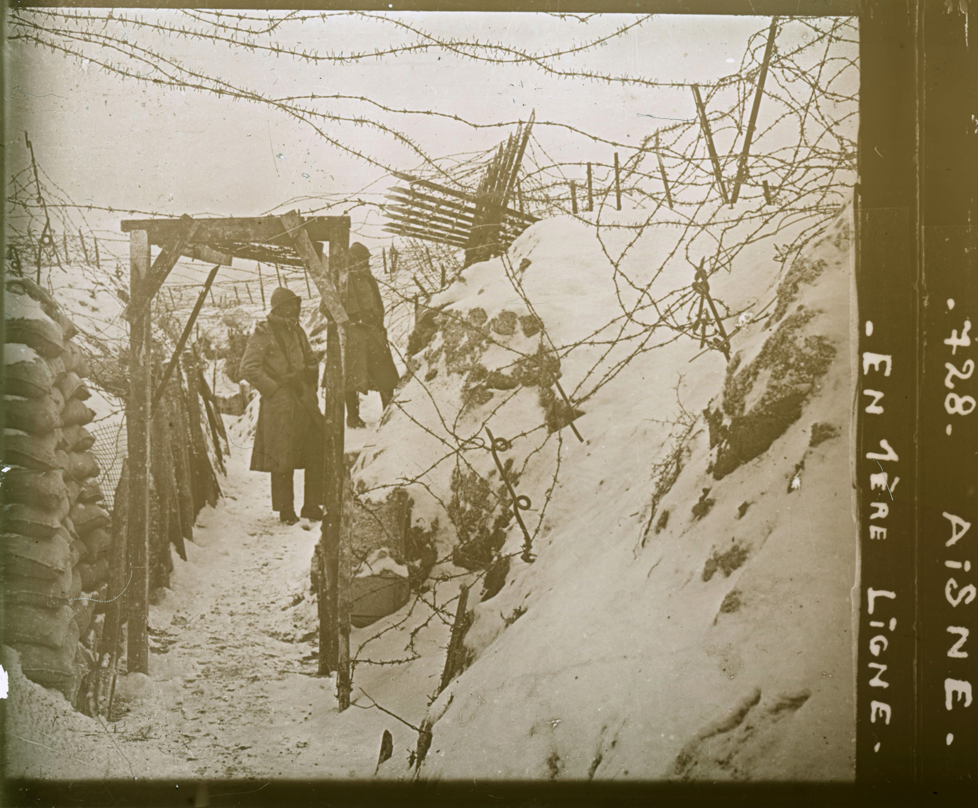 Photographie en noir et blanc d'une tranchée enneigée. Une personne vêtue d'un long manteau se tient debout dans la tranchée, sous un échafaudage et des barbelés.