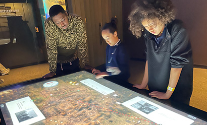 Three young Black learners leaning over a touch screen table with a battlefield scene on it
