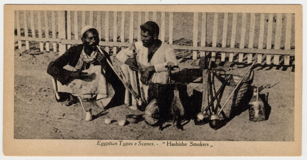Black and white photograph of two men sitting in front of a fence smoking long pipes