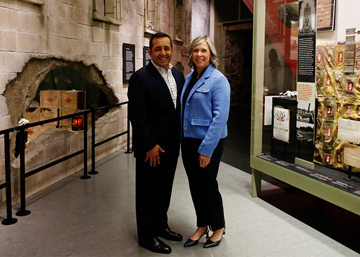 Modern photograph of a brown man in black slacks and jacket, and a white woman in black slacks and a blue jacket, posed in front of the field hospital exhibit in the Main Gallery