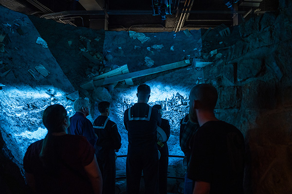 Museum guests stand inside a replica of a devastating bomb crater bathed in blue light as films are projected on the sides of the crater