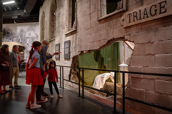 Several young guests look at a museum exhibit depicting a bombed-out cathedral. Nurse and soldier mannequins are visible inside the exhibit