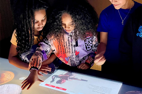 Two Black girls and a white boy crowd around a large touchscreen table, interacting with the graphics