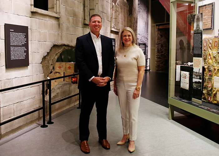 Modern photograph of a tall white man in a black suit and a white woman in cream slacks and sweater posed in front of the field hospital exhibit in the Main Gallery