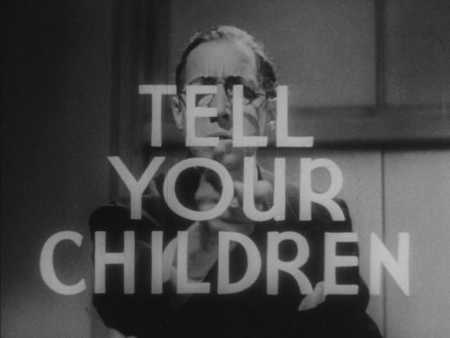 Black and white film still of a white man in a suit facing the camera and pointing at the viewer. The words 'TELL YOUR CHILDREN' are superimposed over him.