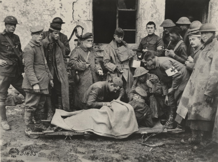 Black and white photograph of a group of soldiers gathered around a chaplain kneeling next to a wounded soldier on a stretcher.