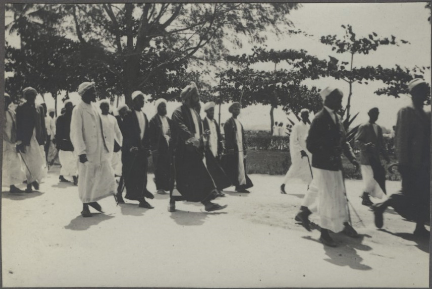 Black and white photograph of a group of Black men in robes and head coverings walking down a tree-lined road