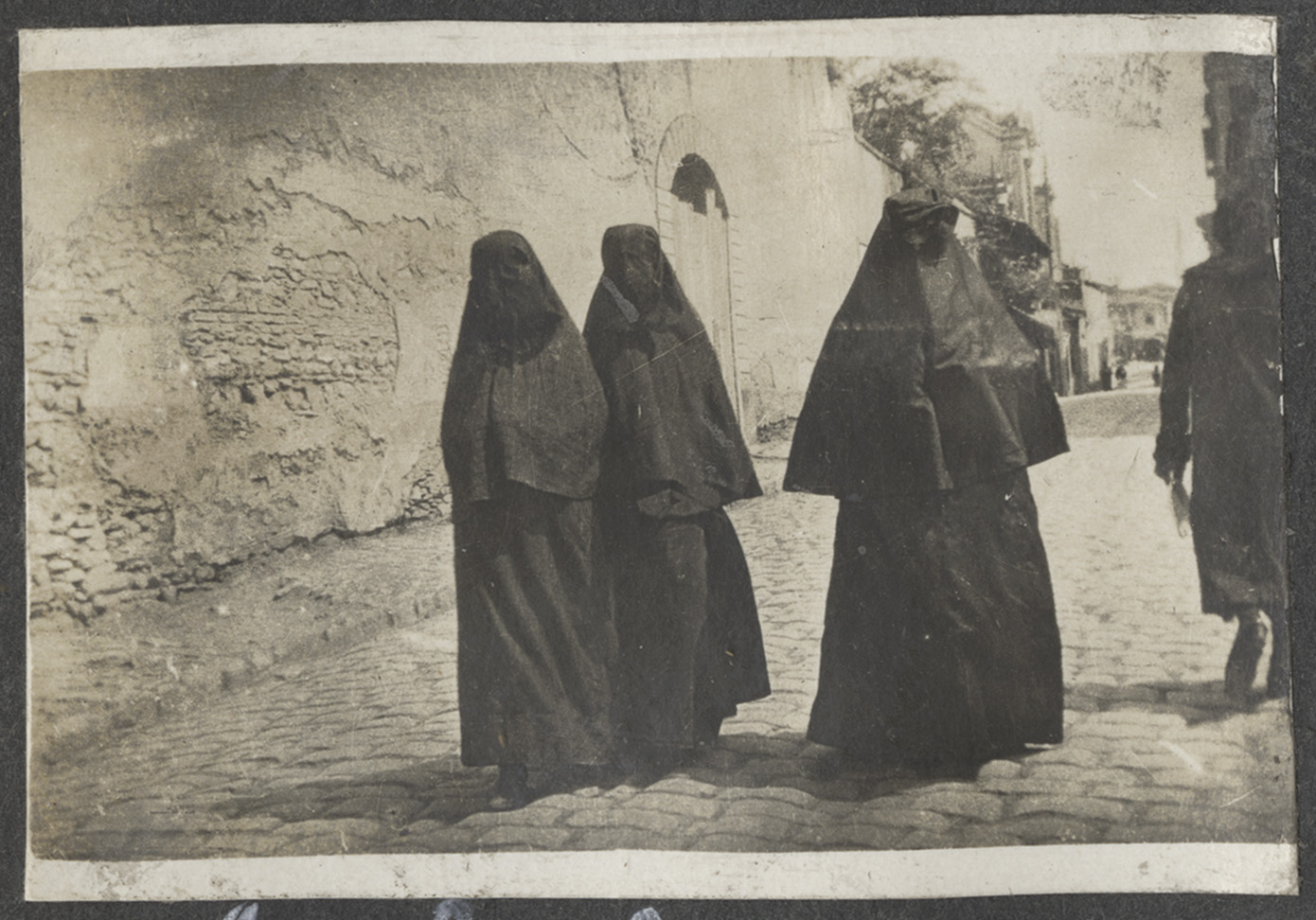 Black and white photograph of three dark-robed and veiled figures walking down a cobblestoned street