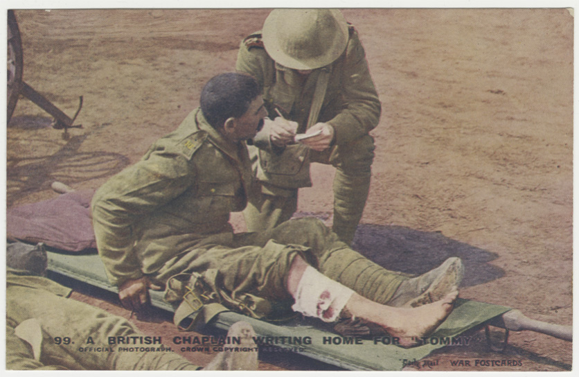 Color photograph of a chaplain in uniform kneeling next to a stretcher with a wounded soldier lying on it. The chaplain is writing on a piece of paper.