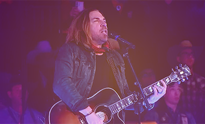 Middle-aged white man with long brown hair wearing a denim jacket and playing an acoustic guitar on a stage while singing into a microphone