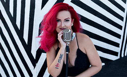 Pale-skinned woman with dyed red hair smiling impishly over an old-fashioned studio microphone