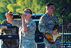 Two men and a woman in camo uniforms play instruments and sing on an outdoor stage