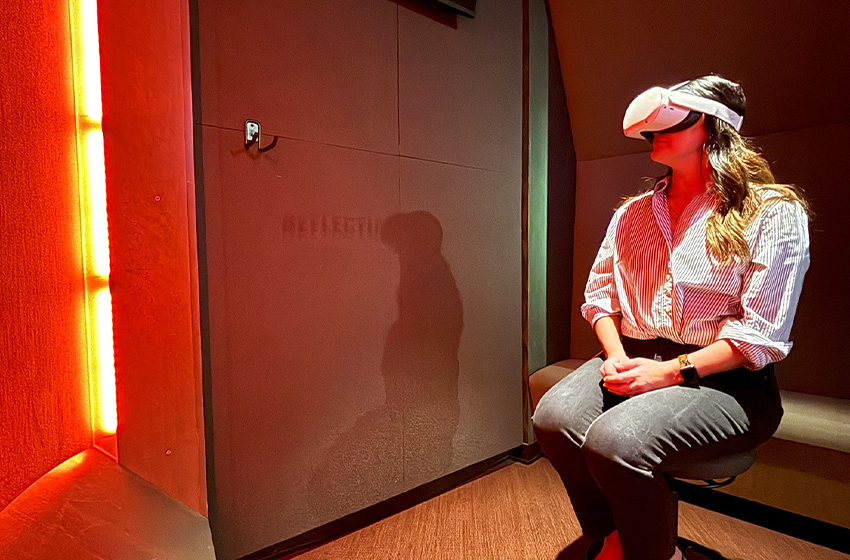 Modern photograph of an enclosed alcove in a museum gallery. A white woman with long brown hair wearing jeans and a button-down sits on a comfortable bench, wearing a large VR headset over her eyes.