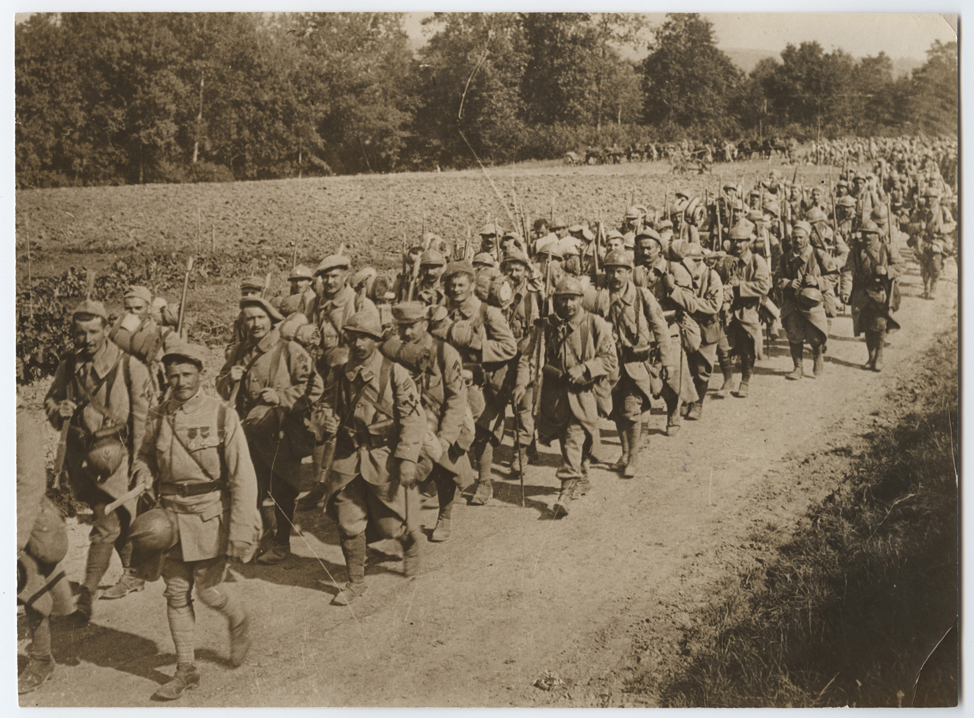 Sepia photograph of a column of WWI soldiers marching on a path between fields