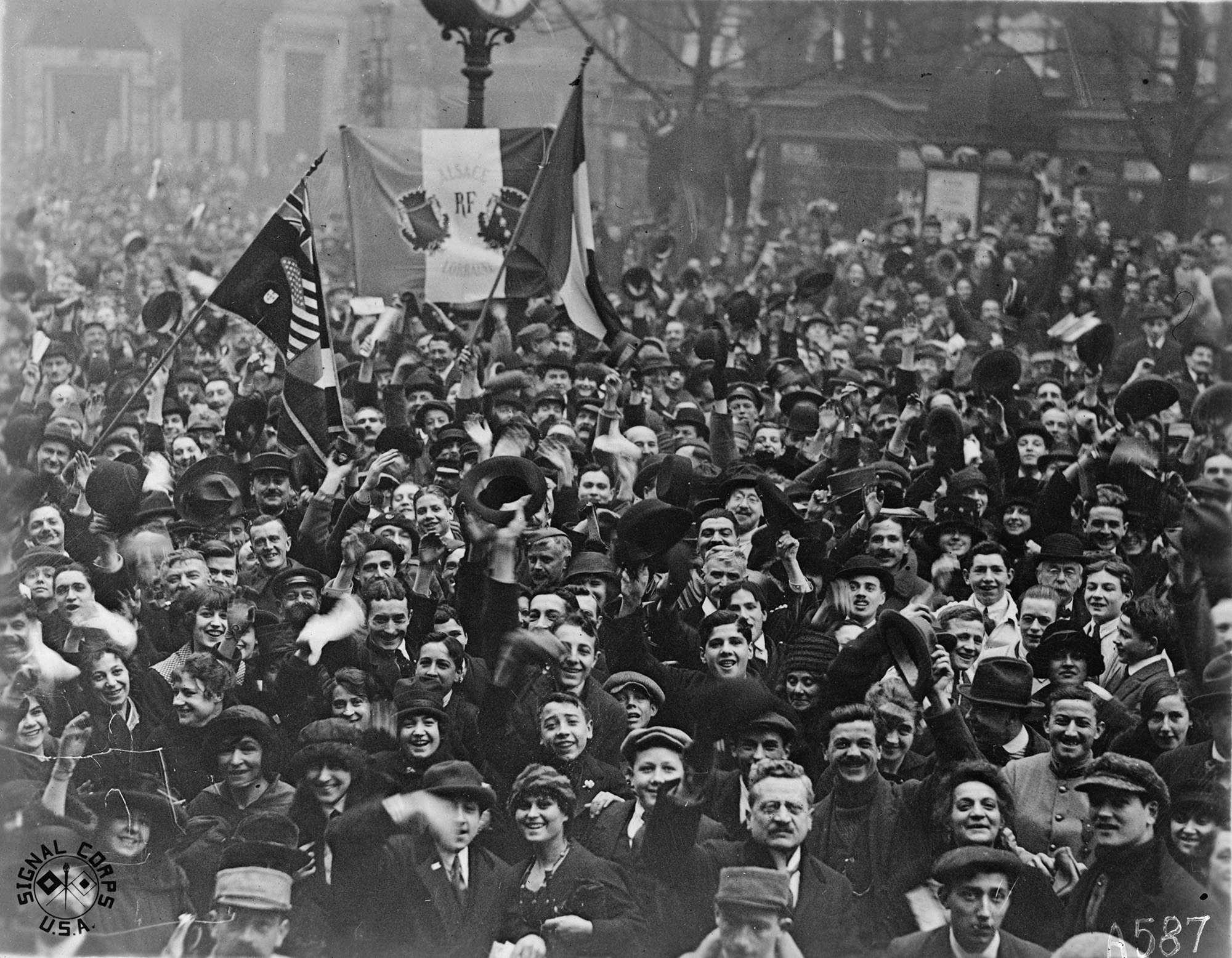 Black and white photograph of a street packed with people smiling and waving flags