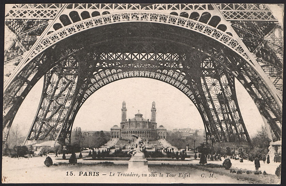Black and white historical photo taken through the base of the Eiffel Tower towards a palace in the distance