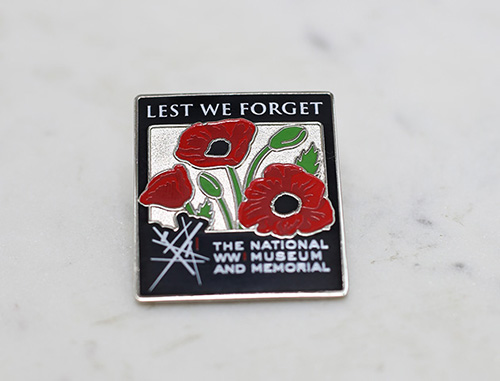 Modern photo of a pin engraved with poppies and the phrase 'Lest We Forget'