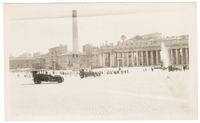 Photo en noir et blanc d'une grande place pavée avec des palais en pierre en arrière-plan et des automobiles et des touristes au premier plan.