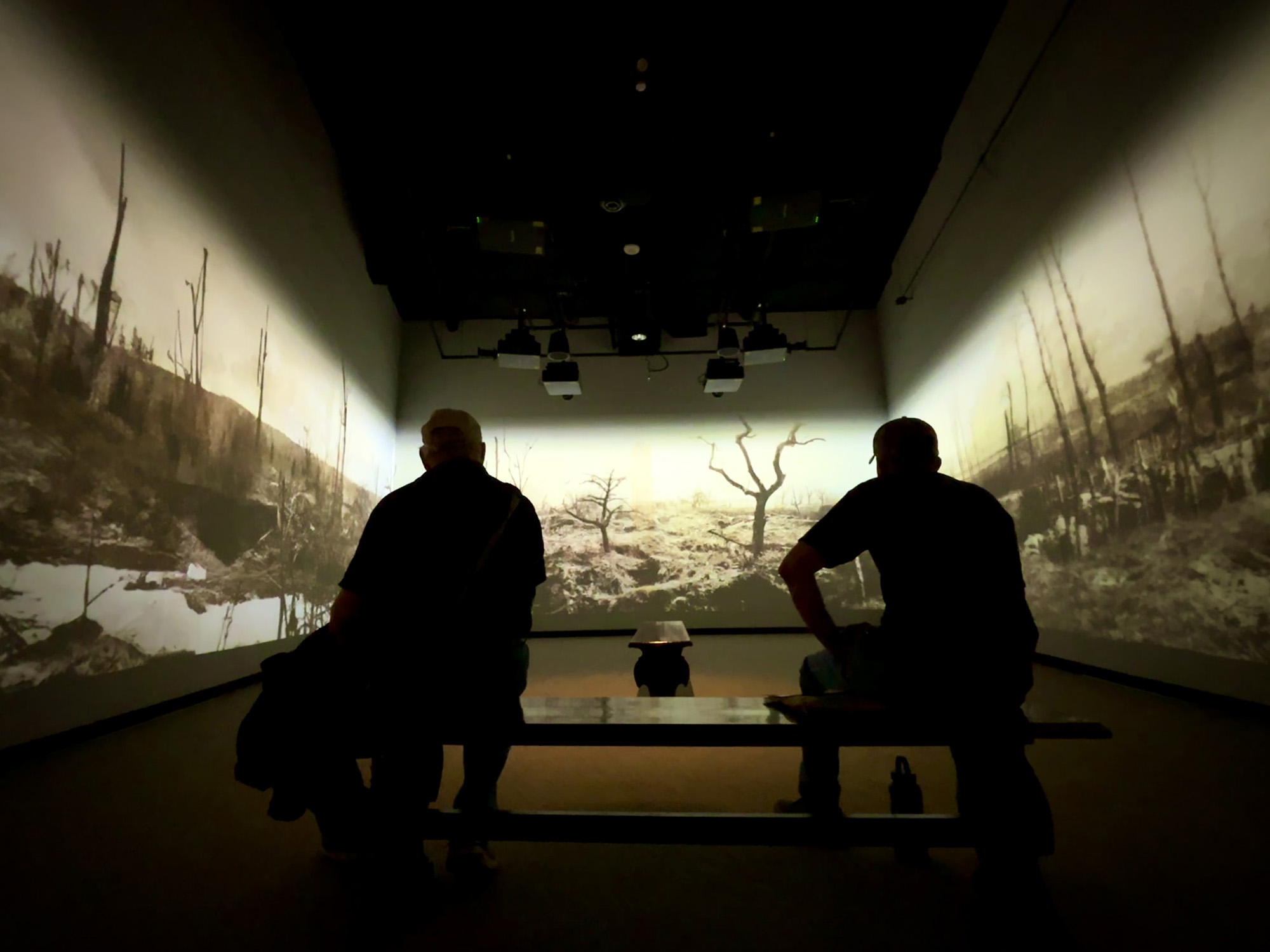 Two guests sit on a bench surrounded by a video projection of a battlefield on three walls