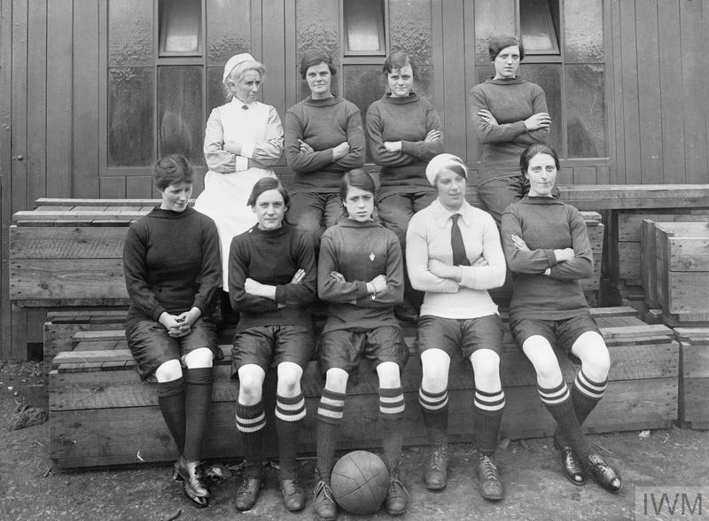 A black and white photograph with five women sitting in the foreground and four women standing behind them. The women are all wearing the same over the knee socks and there is a soccerball at the front center of the image.