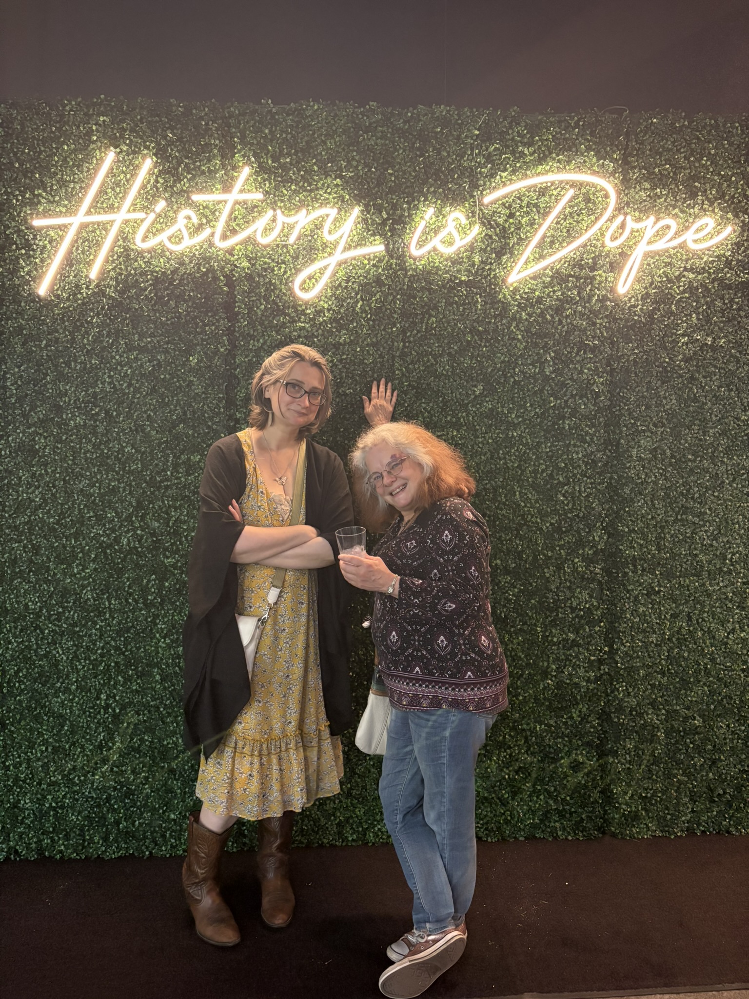 An older white woman and a younger white woman pose in a photo station under a lit neon sign spelling out 'History is Dope'