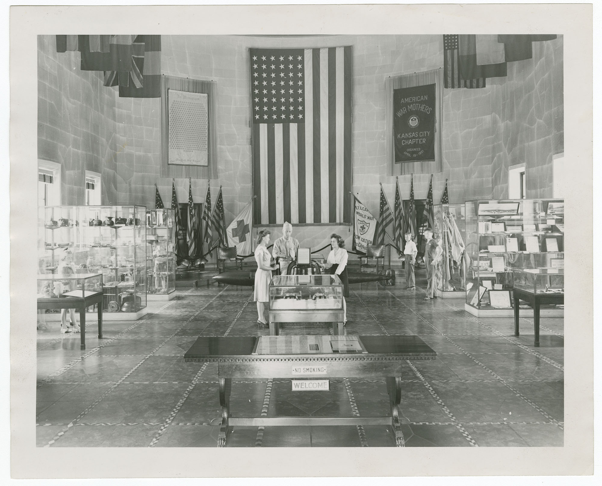 Black and white photograph of the interior of a Museum building. A large American flag is hung at the back of the hall with a flag for the American War Mothers Kansas City Chapter to the right. Glass display cases line each side. Two women and a man in military uniform stand at center.