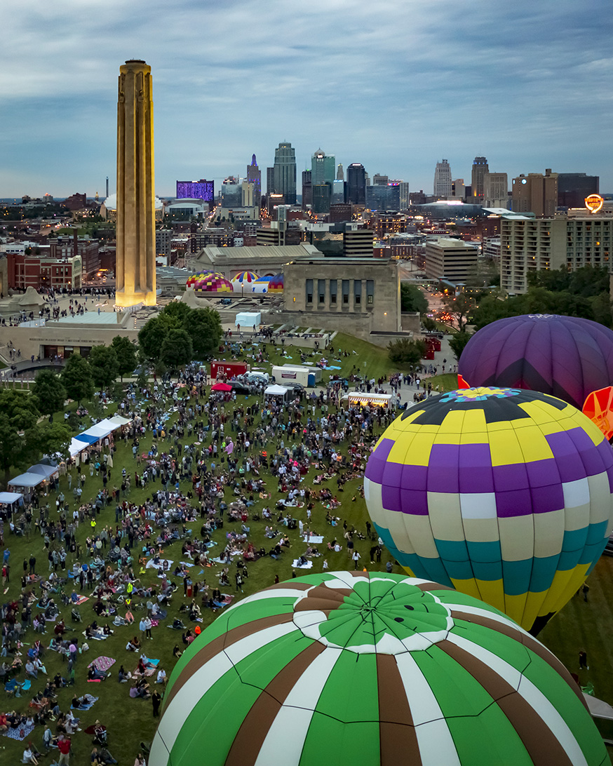 Modern photo of three hot air balloons floating while tethered to the ground. A large crowd of people gather around the balloons. Liberty Memorial Tower in the background.