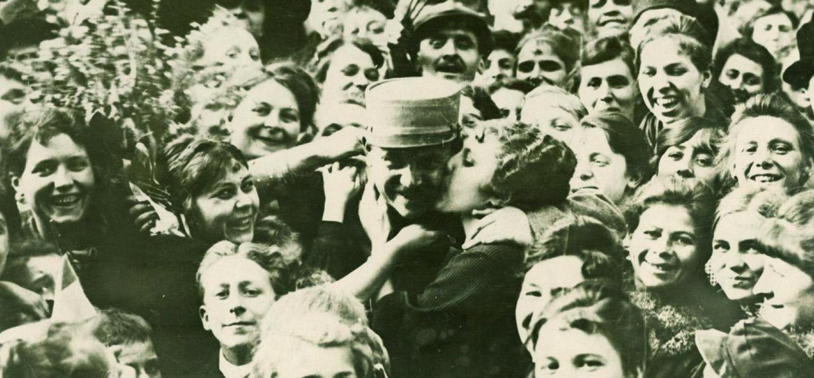 Black and white photograph of a happy crowd. In the center, a woman kisses the cheek of a man in a military cap.