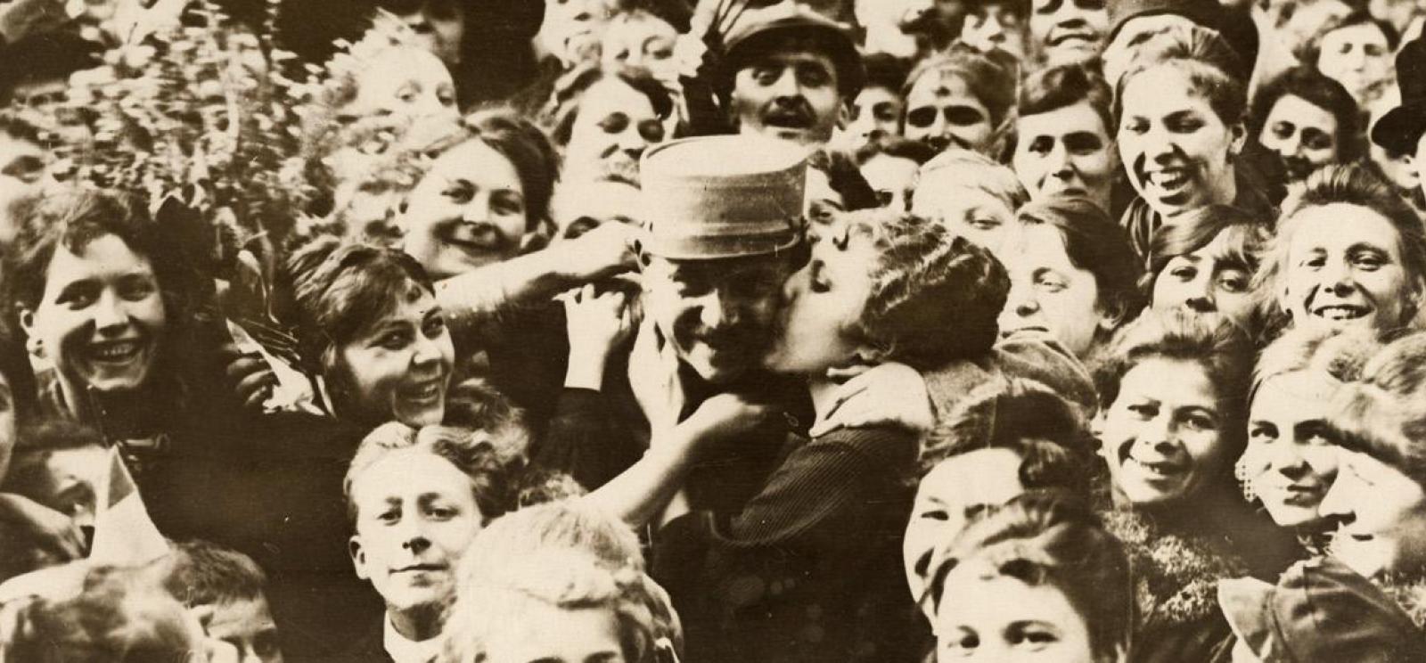 Sepia photograph of a crowd of smiling faces. In the center a woman is kissing the cheek of a man wearing military uniform.