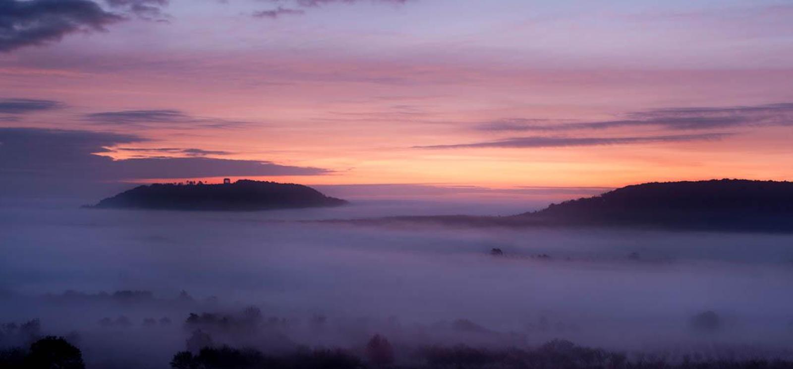 Modern photograph of an empty field and wood at dawn. The ground is covered by a thick mist.