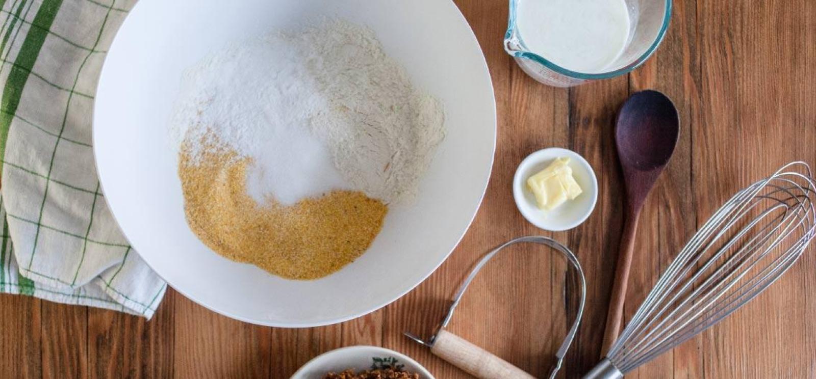 Arrangement of a various baking equipment and supplies: a bowl filled with flour, cornmeal and salt. A small bowl of butter. A measuring cup of milk. A whisk and wooden spoon.