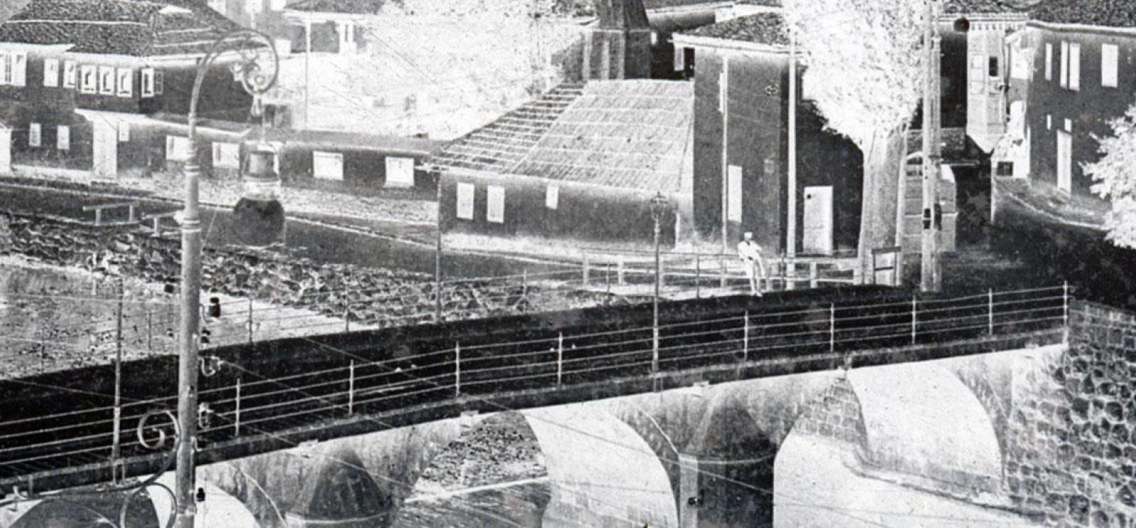 Blackand white photograph with colors inverted so it looks like a photo negative. The image shows a long-distance view of a bridge over a river. The river edge is lined with low houses. There is one person leaning against the railing of the bridge.