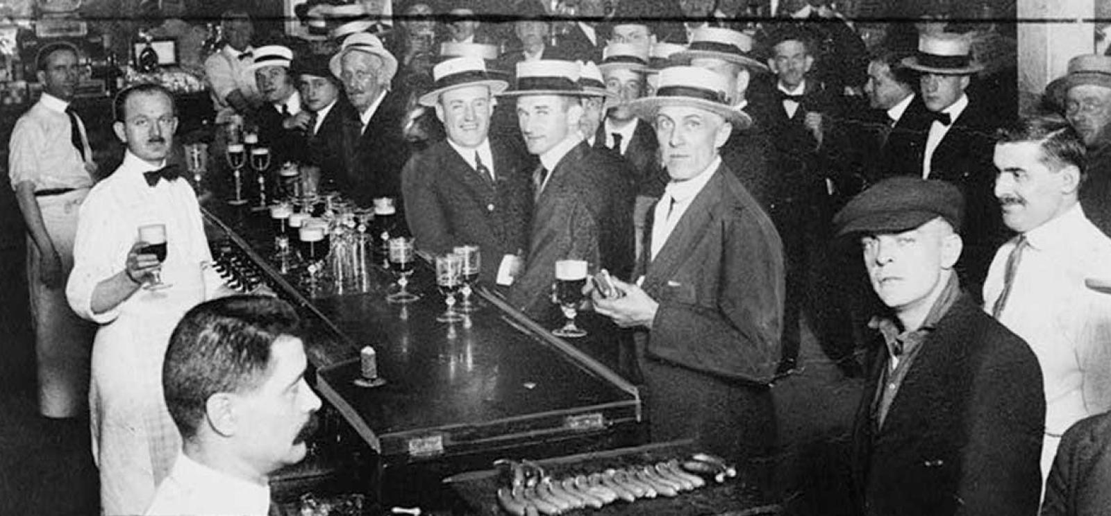 Black and white photograph of a crowded bar with bow-tied bartenders and customers wearing suits and straw boater hats.