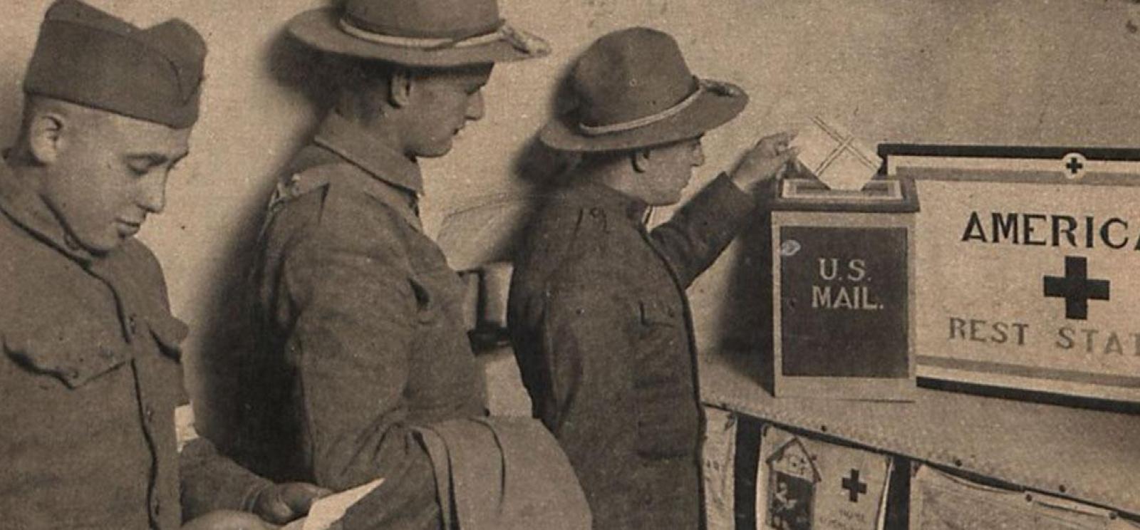 Black and white photograph of three men in military uniform lined up in front of a box labeled 'U.S. Mail'. The one at the front of the line is inserting an envelope into the box.