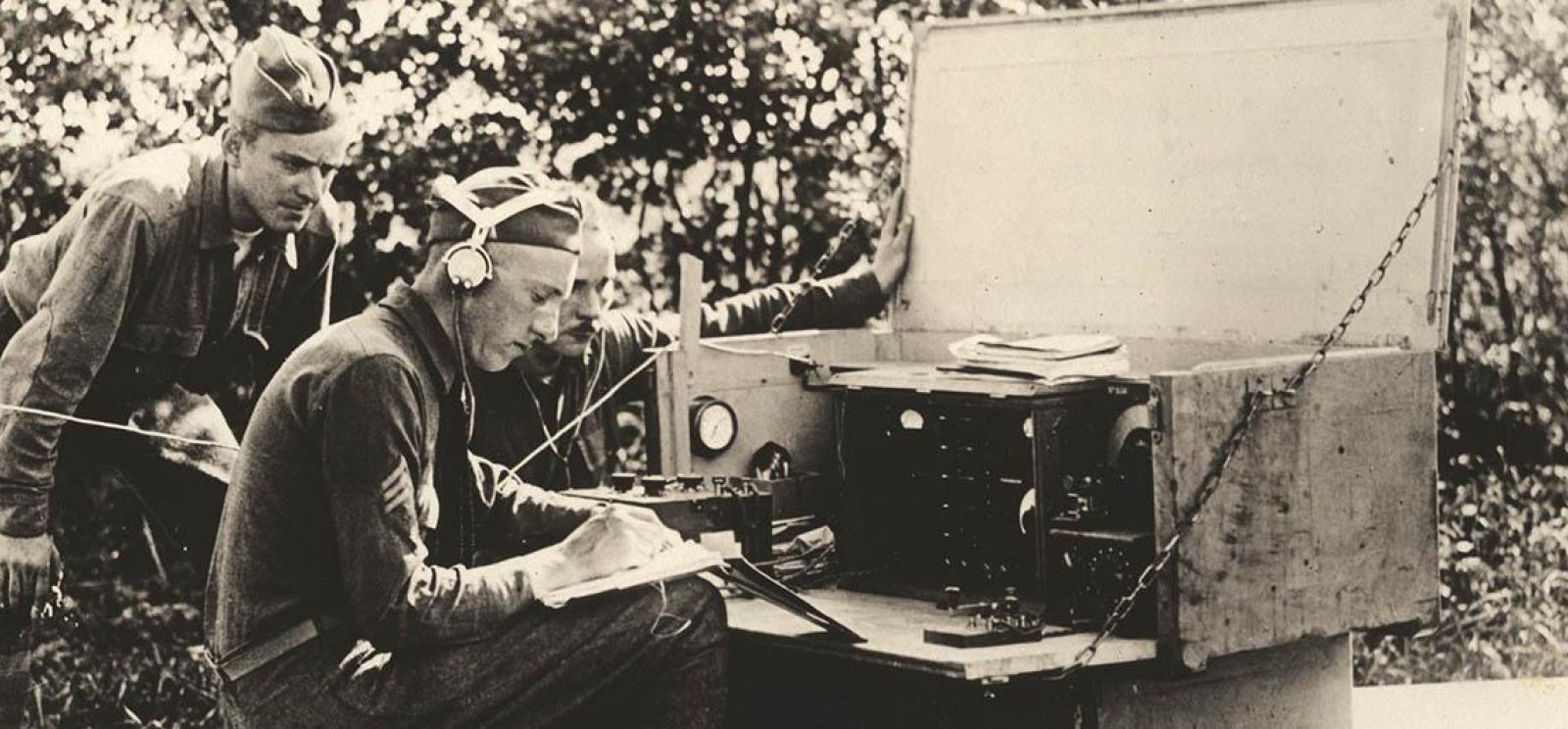 Black and white photograph of three soldiers gathered around a large field radio.