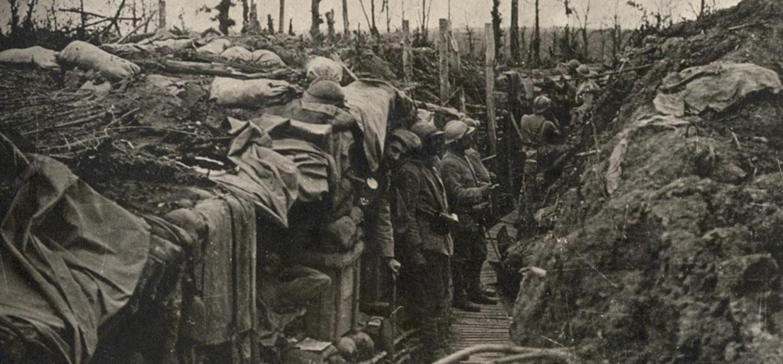 Black and white photograph of a narrow trench. One side is lined with wooden structures. Several men in military combat gear peek out from under the eaves of the structures or stand next to them.