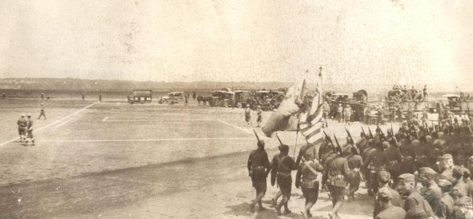 Sepia photograph of a WWI-era baseball game. Soldiers sit in the stands and march by the field.