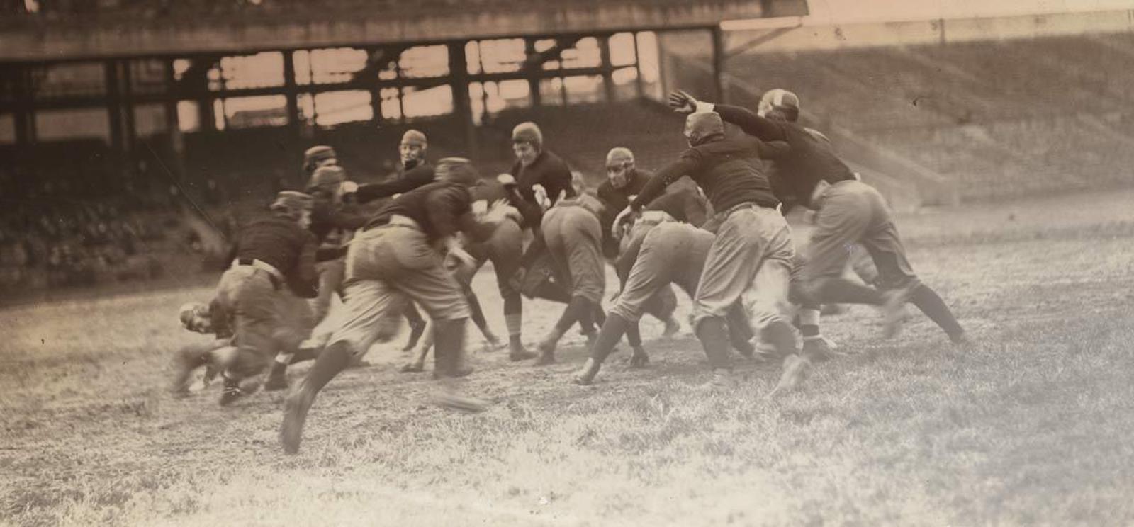Black and white action shot of a group of North American football players mid-play.