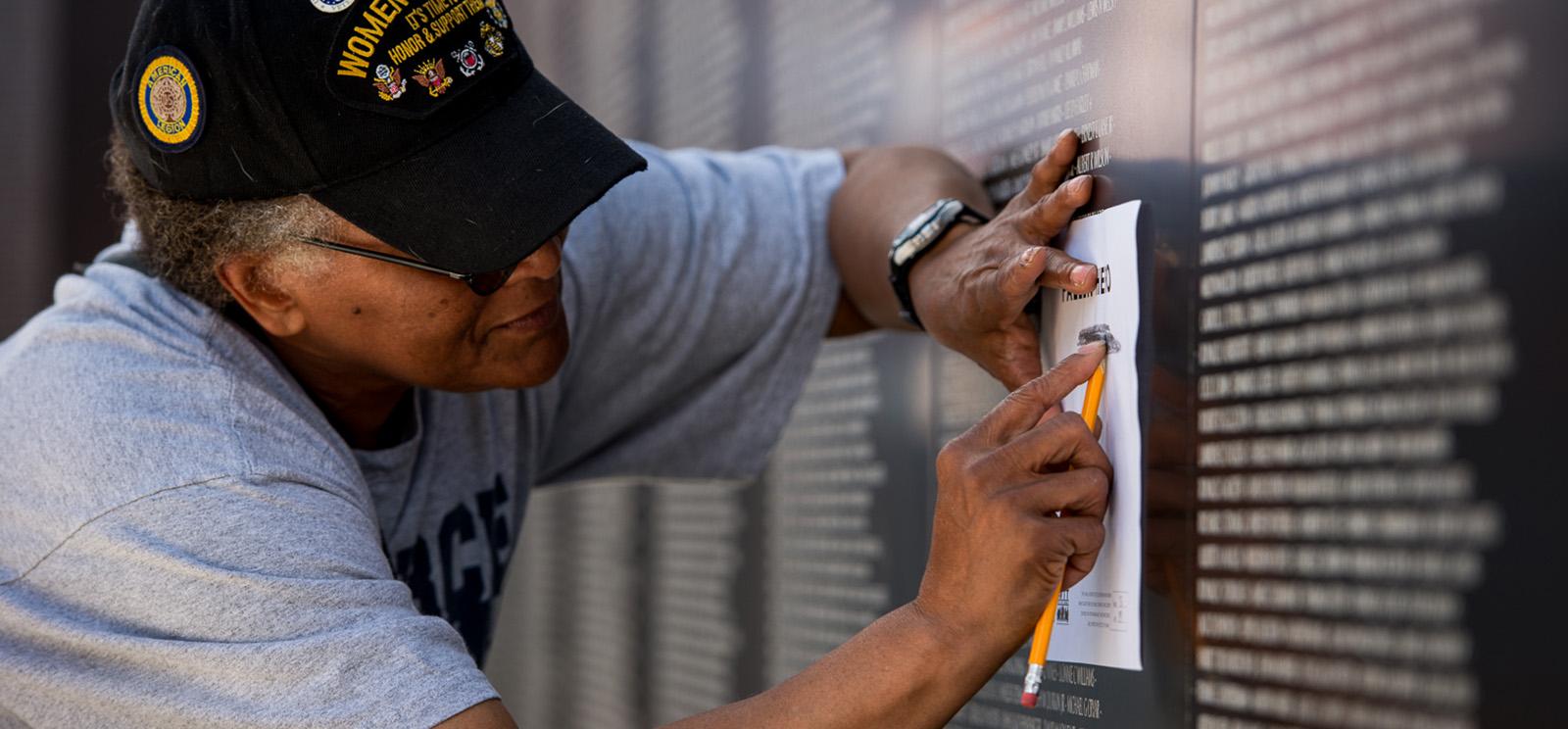 An older Black woman wearing a Woman Veterans baseball cap holds a piece of paper to the etched names on a black wall and creates a rubbing with a pencil.