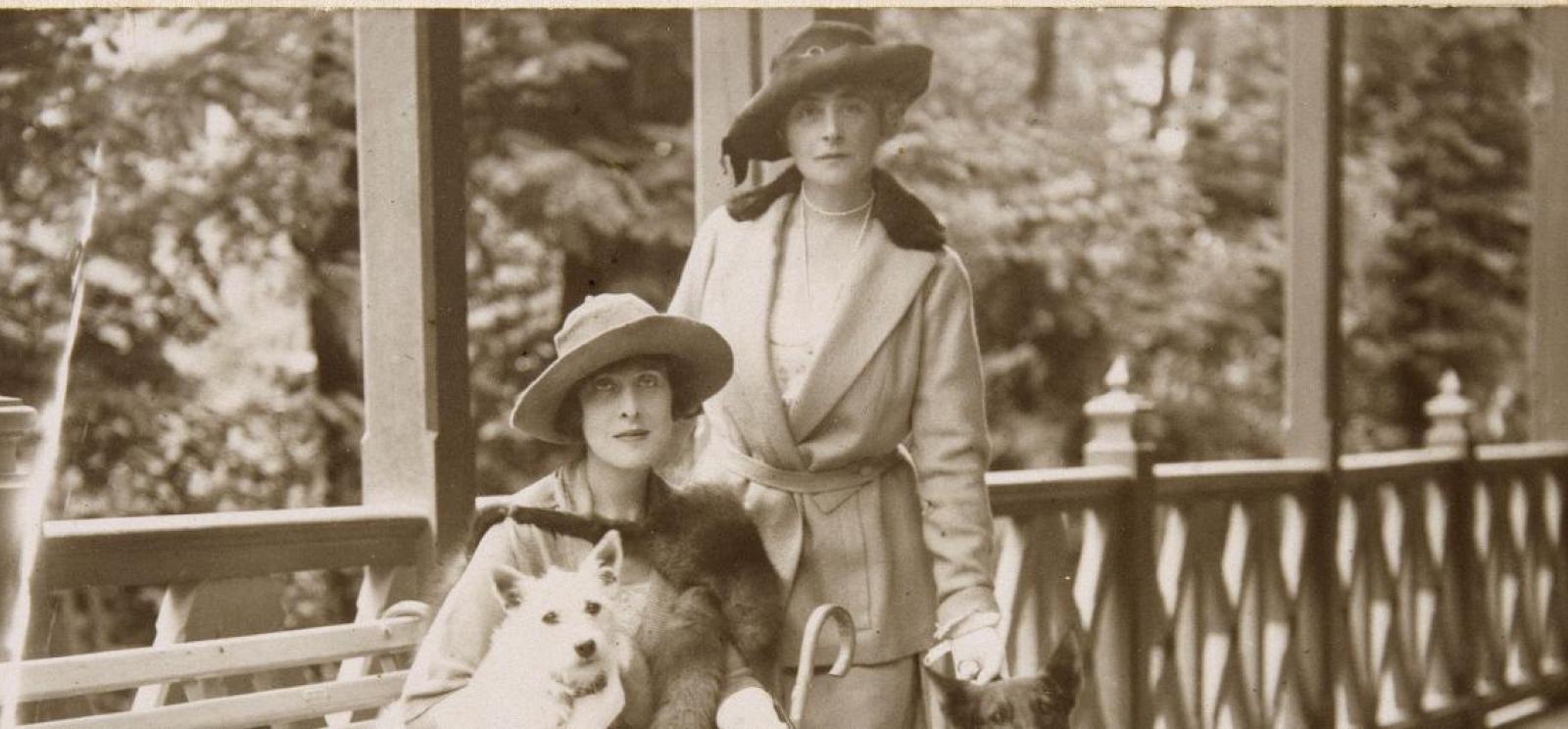 Sepia photograph of two women dressed in walking suits and hats on a building's porch. One is seated with a small dog in her lap.