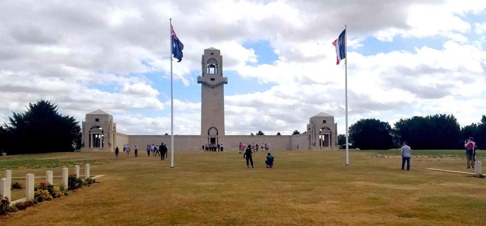 Modern photograph of an open field with a stone war memorial on one end. The memorial features two small buildings on either side and a bell tower in the center. Various tourists sit and stand close and far away from the memorial.