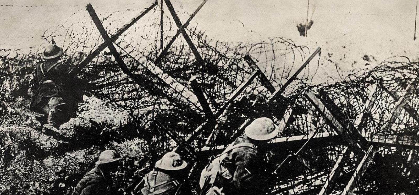 Black and white photograph of a tangle of barbed wire and wooden posts atop a trench through which several soldiers are creeping.