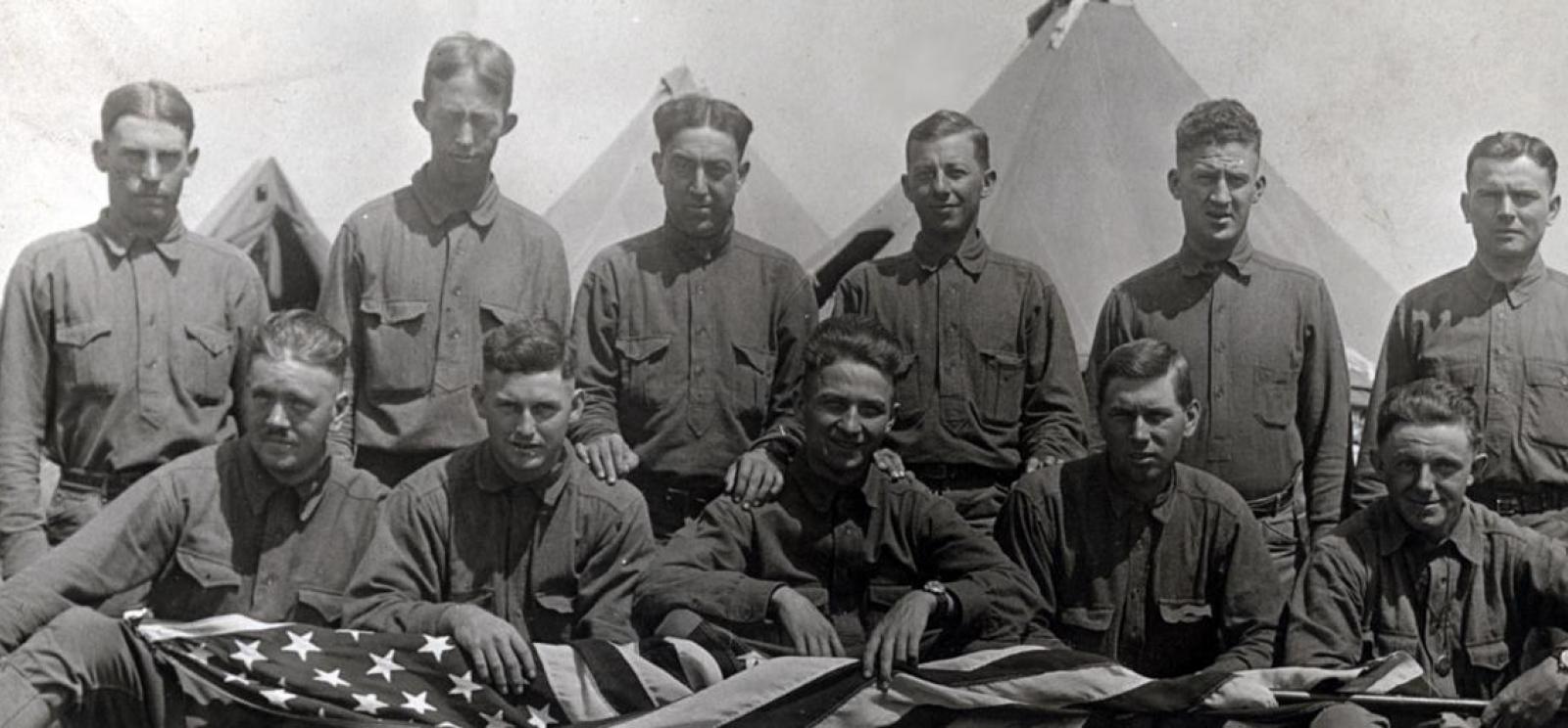 Black and white photograph of a group of soldiers posing in front of tents. The front row holds a U.S. flag. 