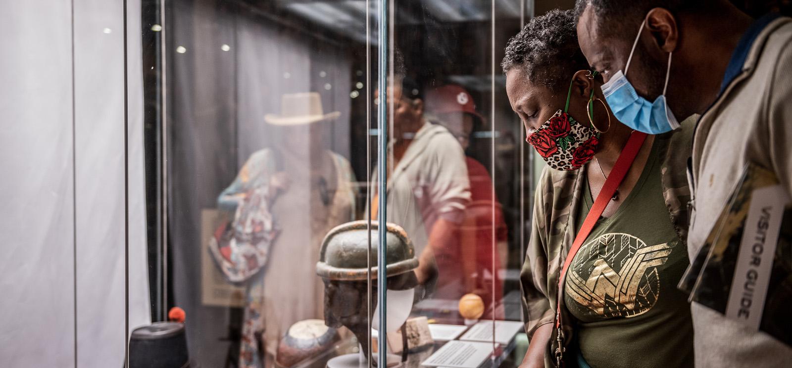 Modern photograph of a Black woman and a Black man wearing masks looking at a glass museum display case of artifacts.