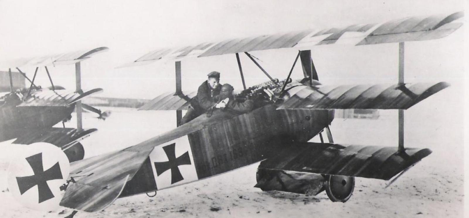 Black and white photo of a WWI-era plane with three layers of wings. A soldier is perched near the cockpit conversing or helping the pilot. The tail and side are decorated with an iron cross.